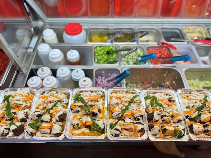 Falafel dishes in trays, with various toppings, next to a counter of salad fixings and sauces.