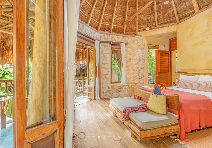 Bedroom with thatched roof, stone walls, and wooden furniture, open to a balcony with greenery.