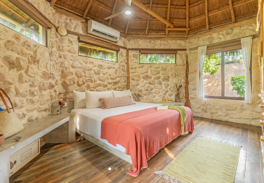 Bedroom with stone walls, thatched roof, and double bed with coral-colored blanket.