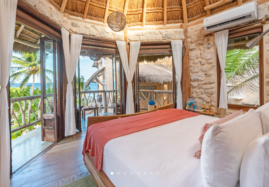 Bedroom with a thatched roof and balcony overlooking the ocean. Light-colored walls, bed with pink throw.