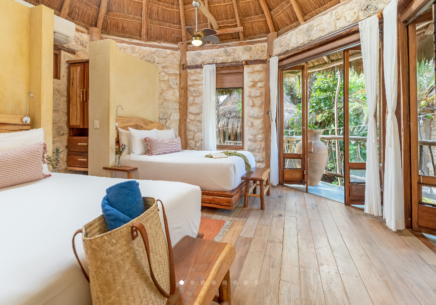 Round room with two beds, open door to balcony, stone walls, wood floors, thatch roof.