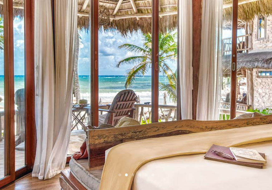 Bedroom with ocean view; wooden bed, deck with chair, palm trees, and turquoise water visible.