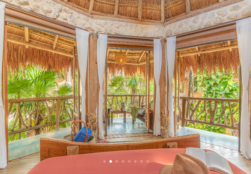 Open-air spa with thatched roof and balcony overlooking lush greenery; red massage bed.