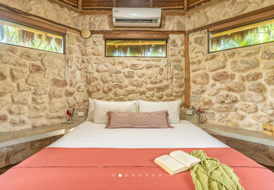 Bedroom with stone walls, double bed, red blanket, open book, and air conditioning unit.