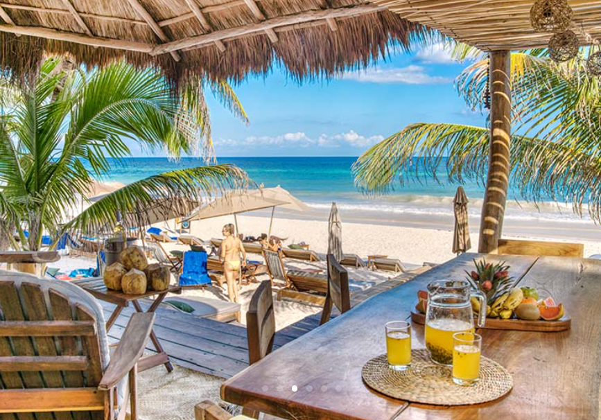 Beach bar with thatch roof overlooking ocean; drinks, coconuts, and people relaxing.