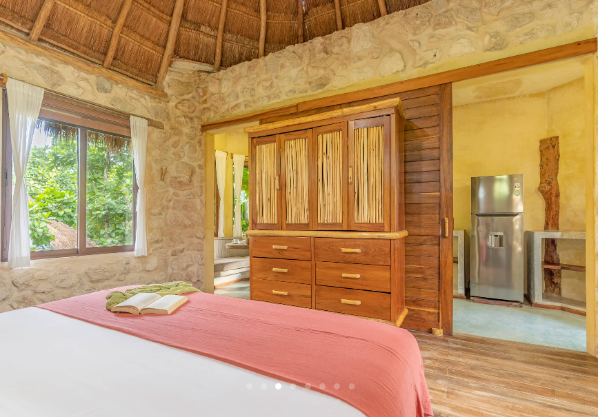 Bedroom with stone walls, thatched roof, wooden wardrobe, bed with pink blanket, and a small kitchen visible.