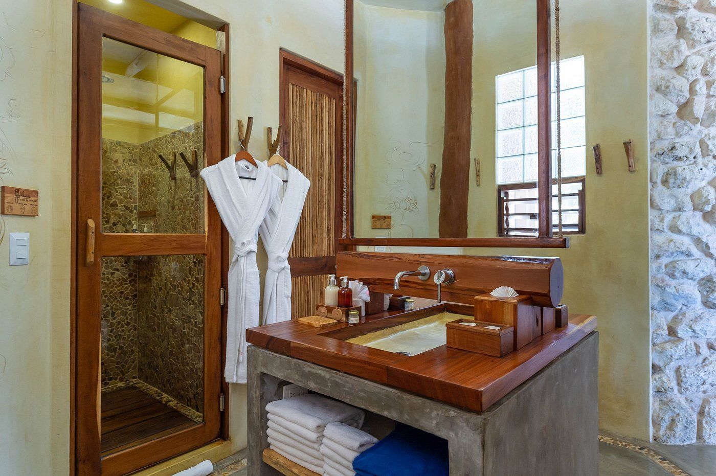 Bathroom with stone walls, wooden accents, and a shower. Robes hang on the door, sink area with toiletries.