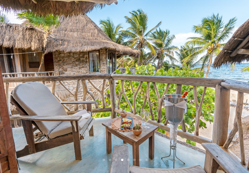 Balcony overlooking beach with chairs, table, champagne bucket, thatched roof buildings, and palm trees.