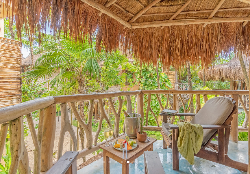 Wooden balcony with chairs, table with snacks, champagne bucket, and lush tropical greenery.