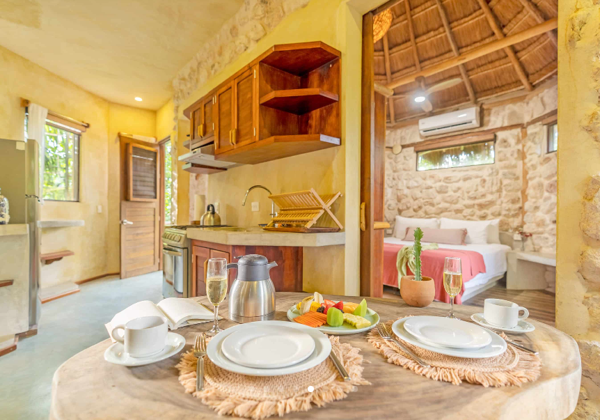 Interior of a round cottage with a dining table set, kitchen, and bed visible; stone walls, thatched roof.