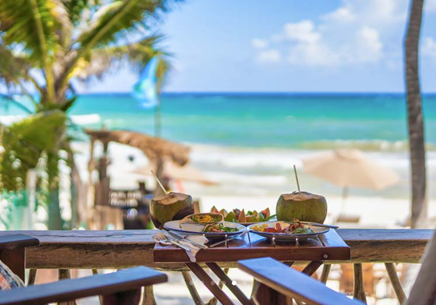 Wooden table with coconuts and fruit, overlooking a tropical beach with turquoise water.