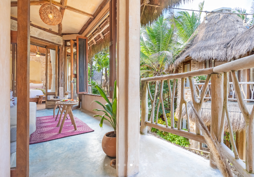 Balcony view of a rustic, tropical-style home with wooden railings, thatch roof, and lush greenery.