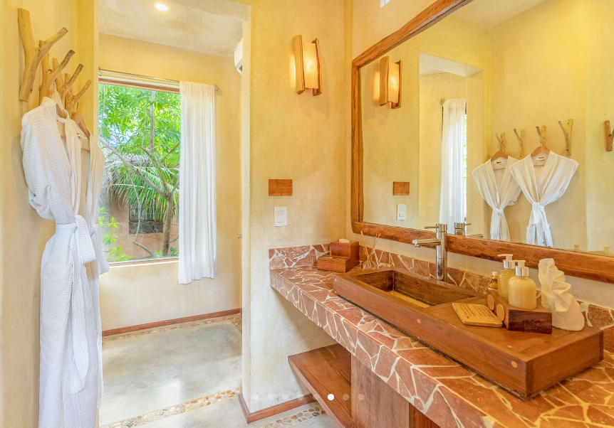 Bathroom with a wooden sink, stone countertop, and two white robes hanging. Window with white curtains.