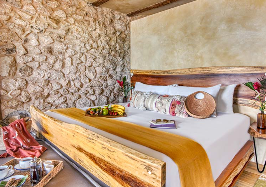 Bedroom with stone wall and wooden bed frame, bed adorned with pillows and an orange runner, with fruit and breakfast.