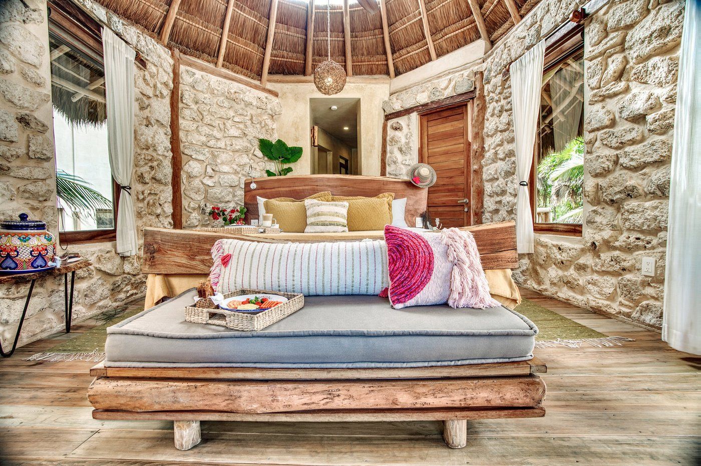 Rustic bedroom with stone walls, wooden bed frame, thatched roof, and window with white curtains.