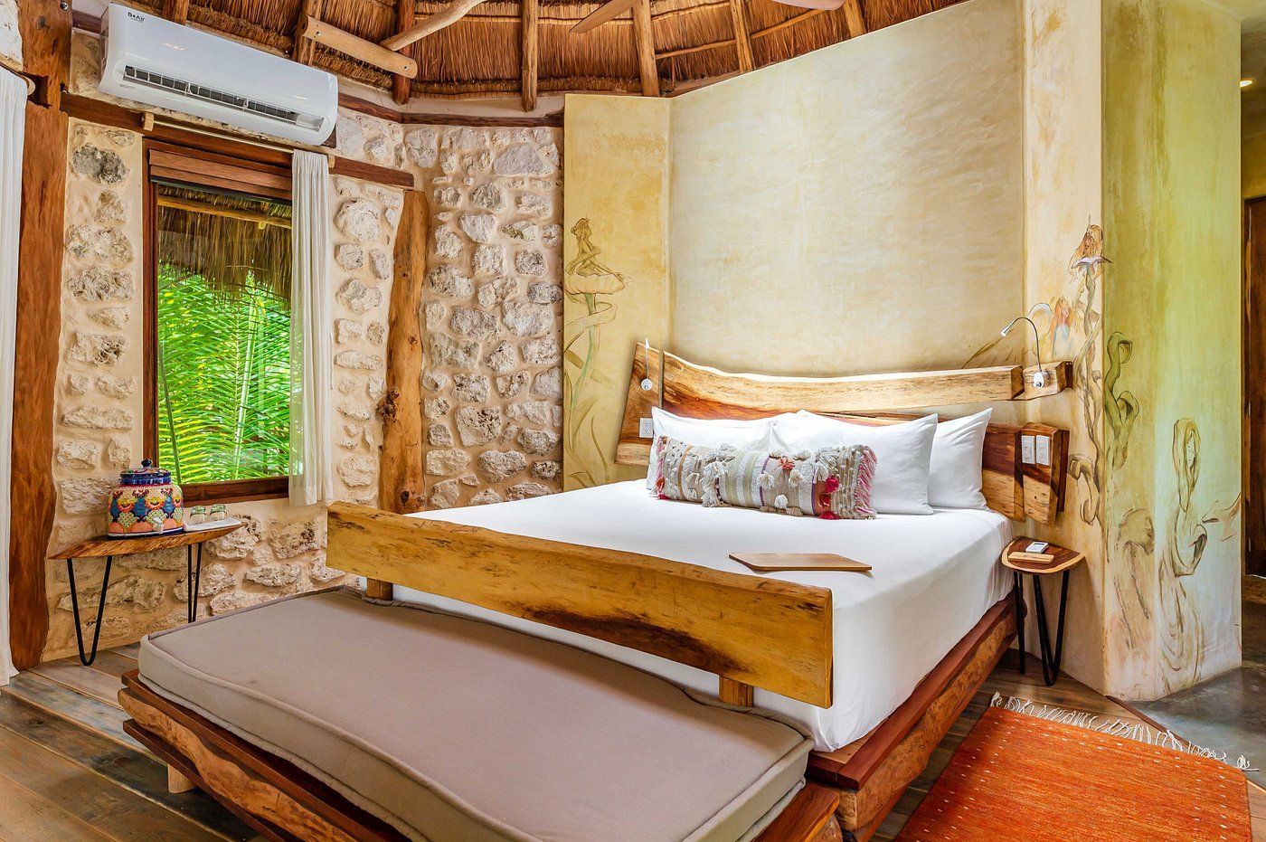 Bedroom with stone walls, wooden bed frame, bench, and window overlooking greenery.