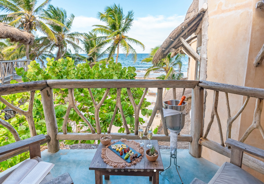 Balcony overlooking ocean, with table of food and drinks. Palm trees and blue sky visible.