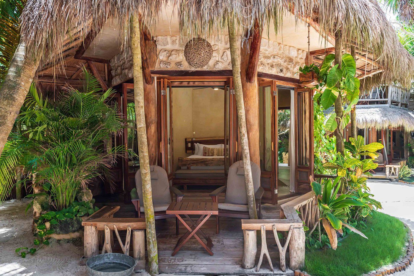 Exterior of a tropical cabana with open doors; two chairs and a table on a porch, thatched roof.