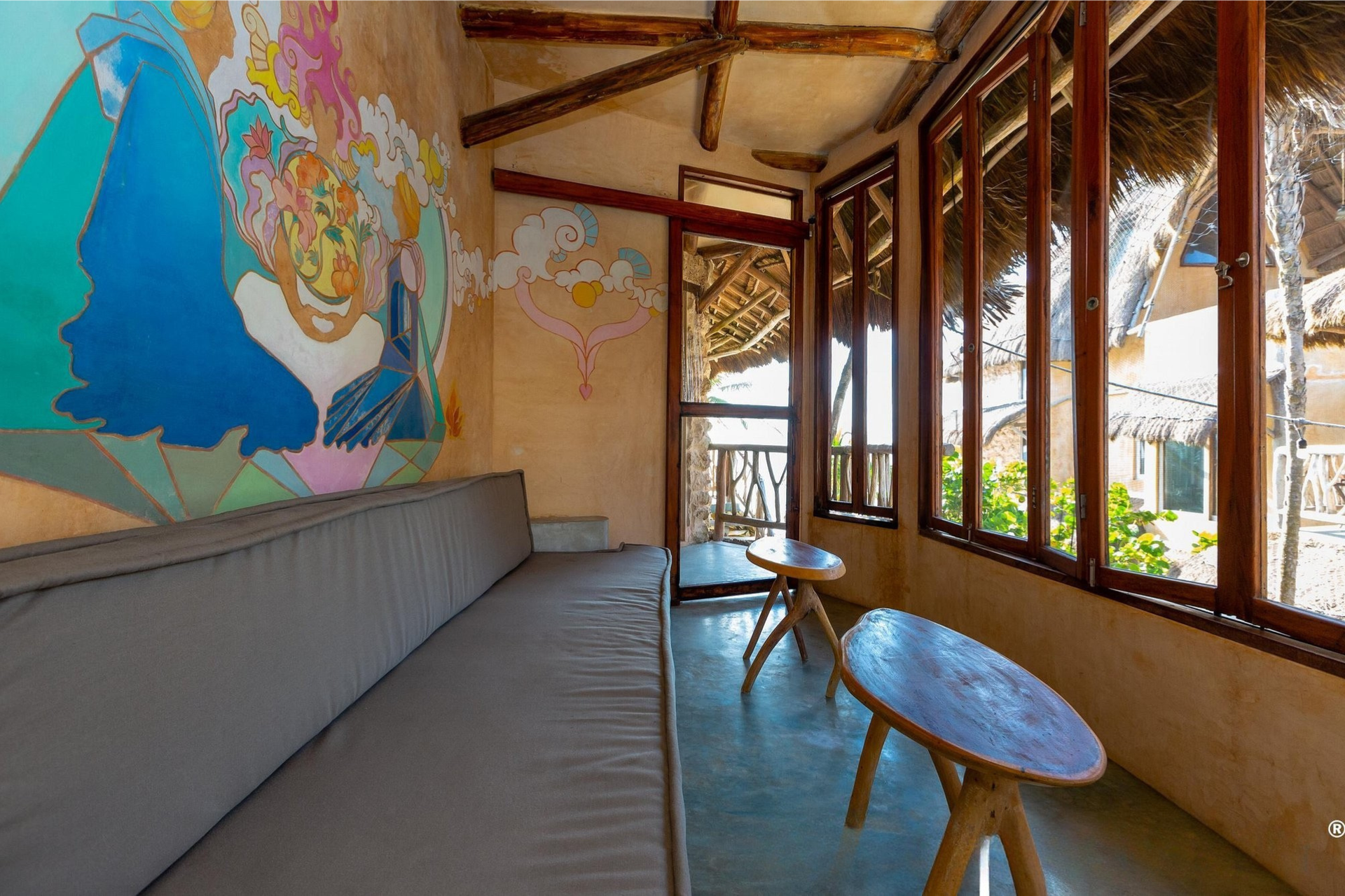 Cozy room with mural, long bench, small wooden tables, and large windows overlooking the outdoors.