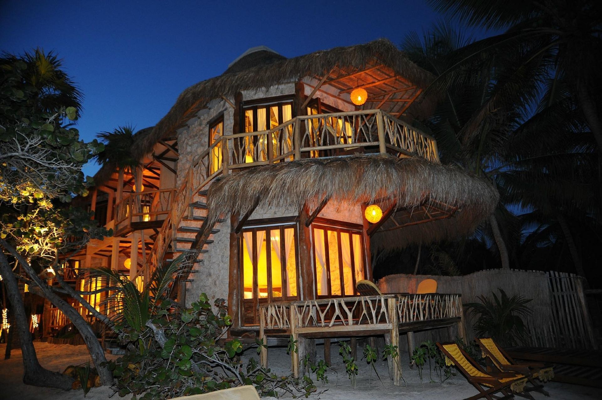 Two-story beach house with thatched roof, lit windows, and wooden deck at night.