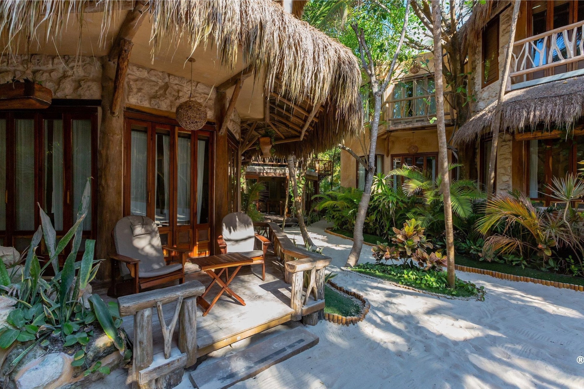 Courtyard with rustic buildings, thatched roofs, wooden furniture, and tropical plants.