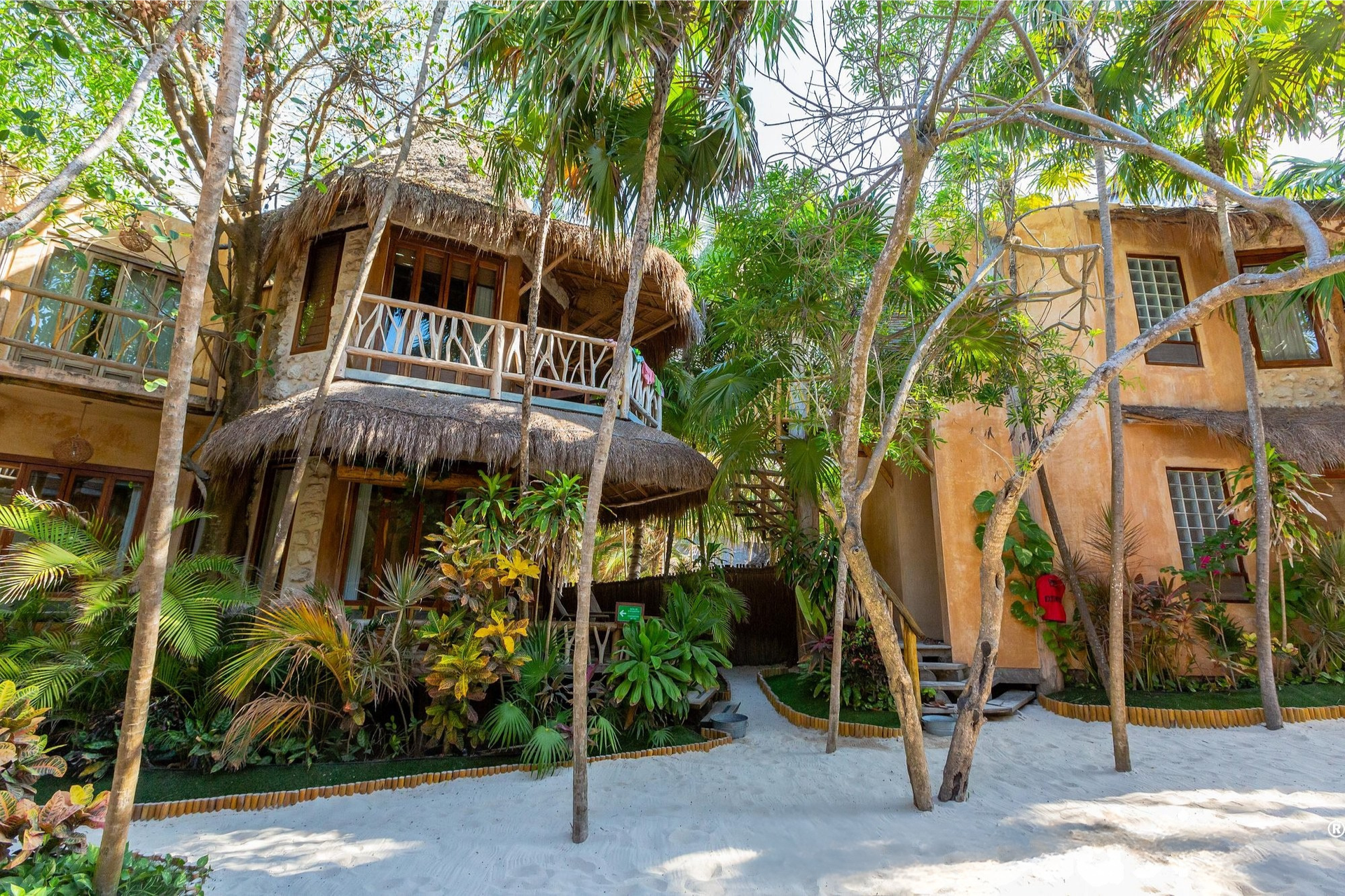 Two-story building with thatched roof and balcony, surrounded by tropical trees and sandy pathway.
