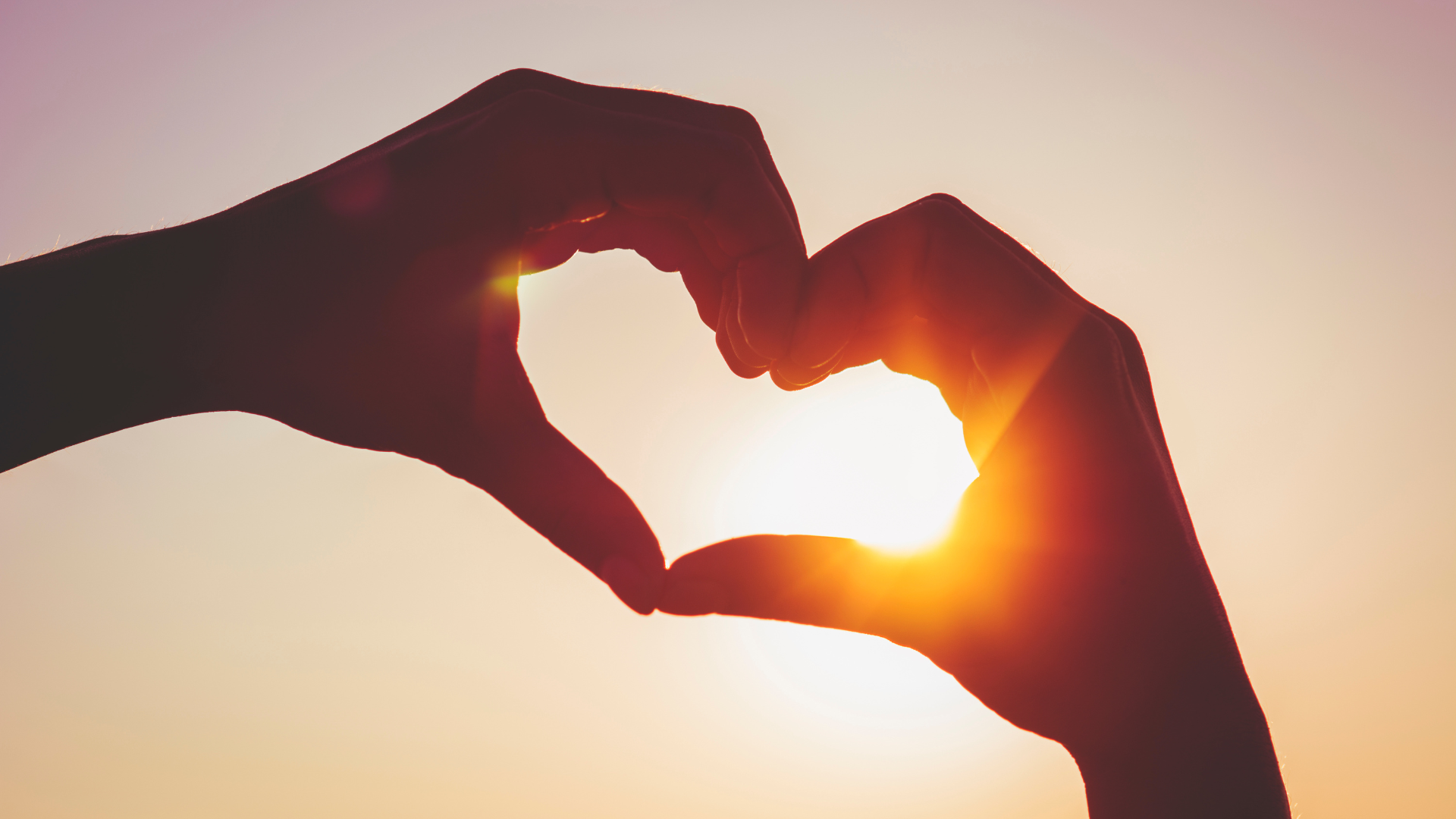 A person is making a heart shape with their hands in front of the sun.