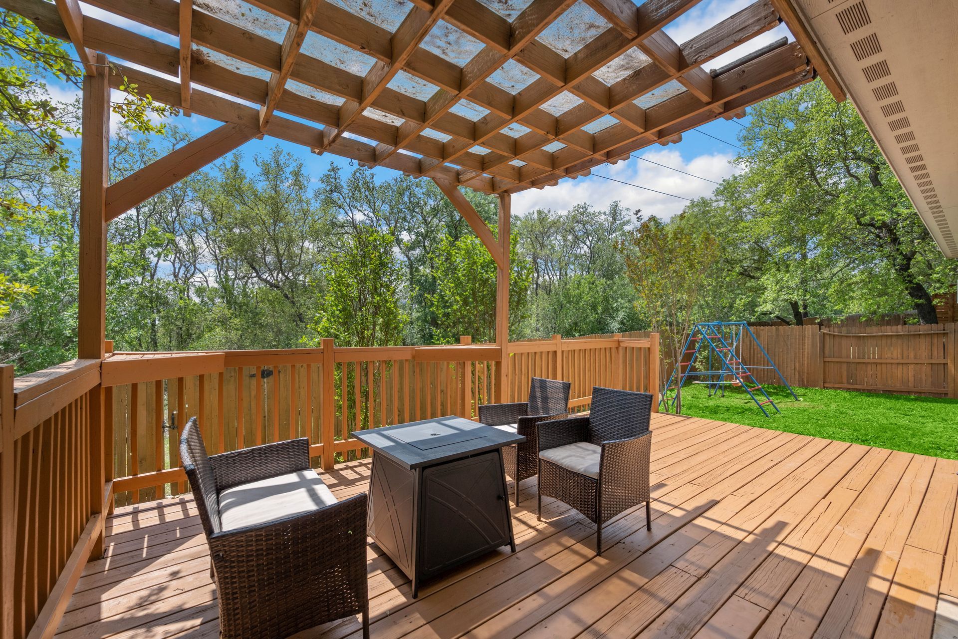 Wooden deck with patio furniture, shaded by a pergola. Lush green trees and grass are in the background.