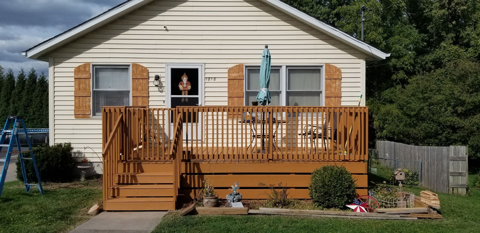 Small house with a brown wooden deck, shutters, and a garden. Green lawn and trees surround it.