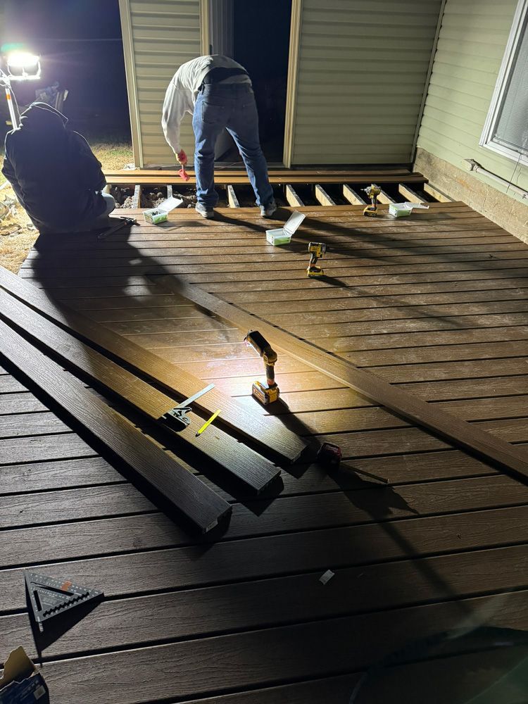 Two people building a brown deck at night using tools; a light shines on the deck.