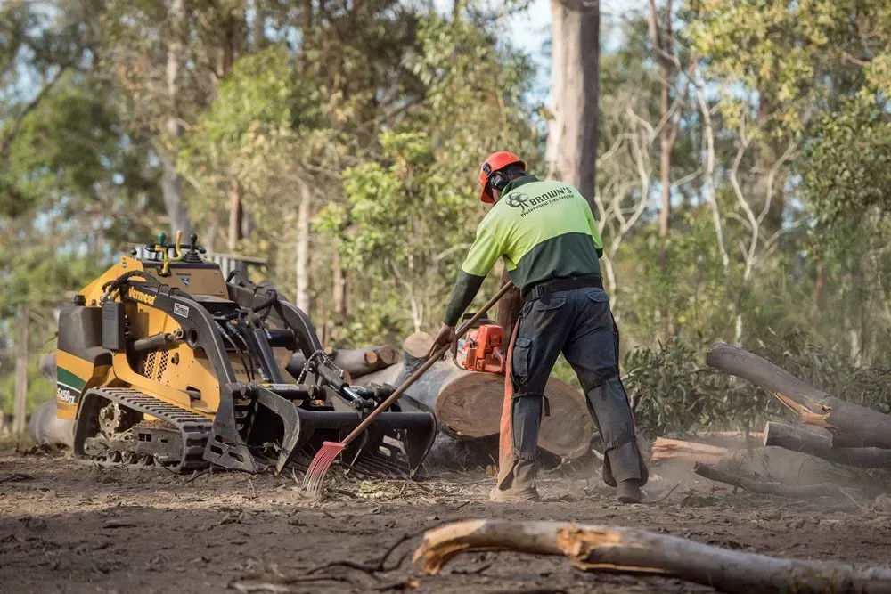 A Person Using a Chainsaw Clears Wood Debris Near a Compact Track — Brown's Professional Tree Service In Pie Creek, QLD
