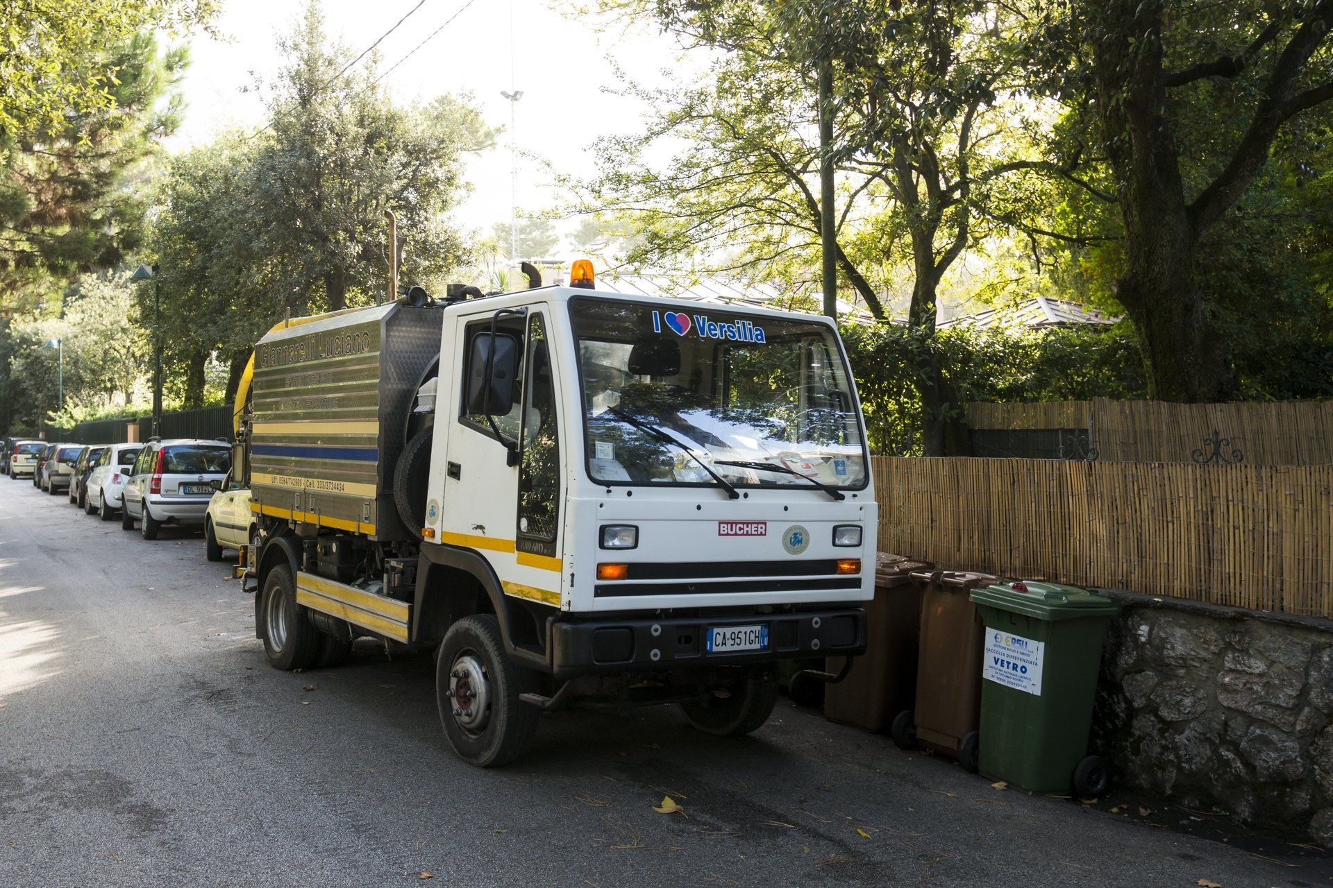 Un camion bianco in una strada stretta, accanto a bidoni verdi e auto parcheggiate. Alberi e fogliame sullo sfondo.