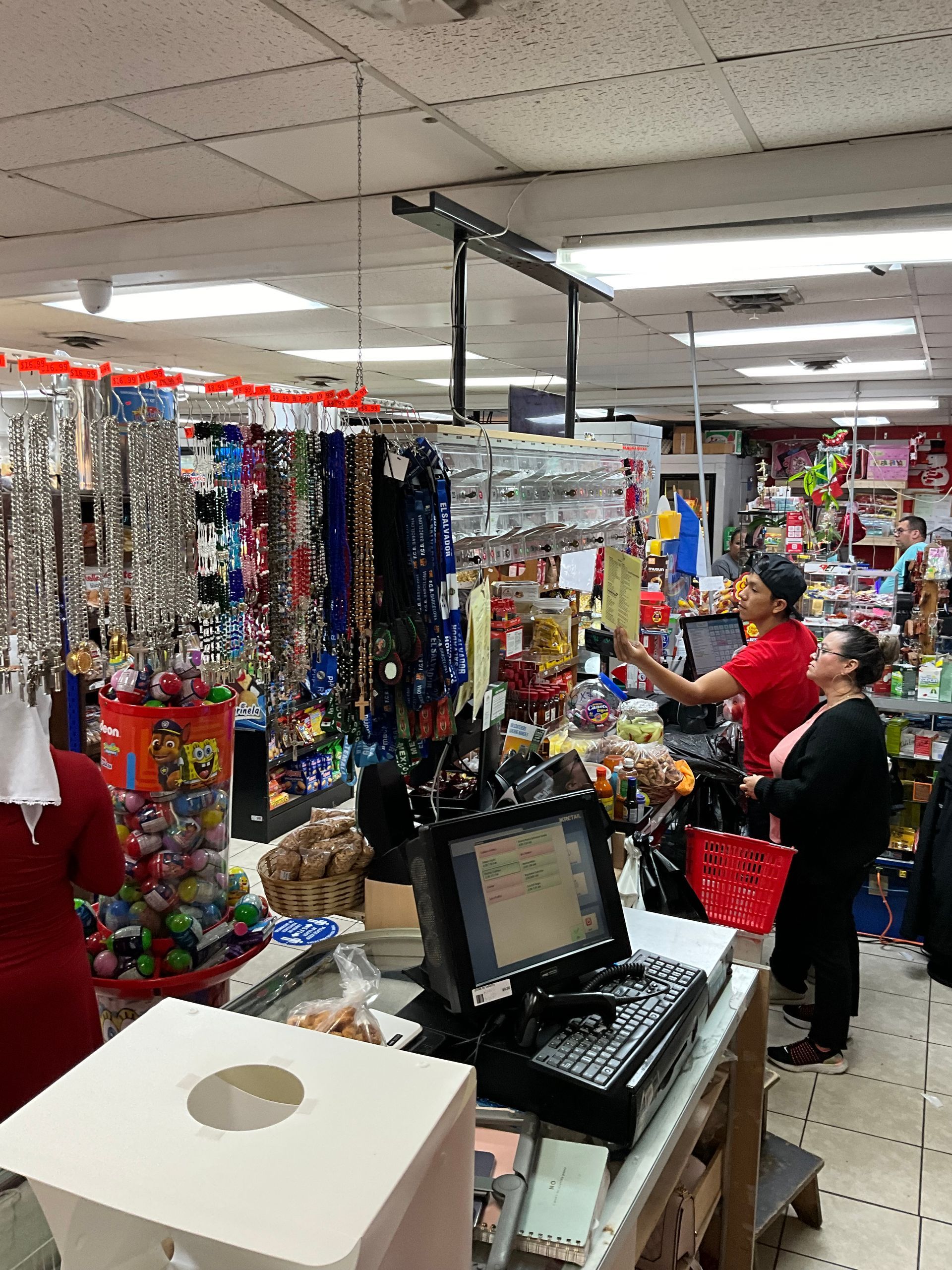 Interior of a store. Two people stand near a cash register, looking at items. Shelves with goods in background.