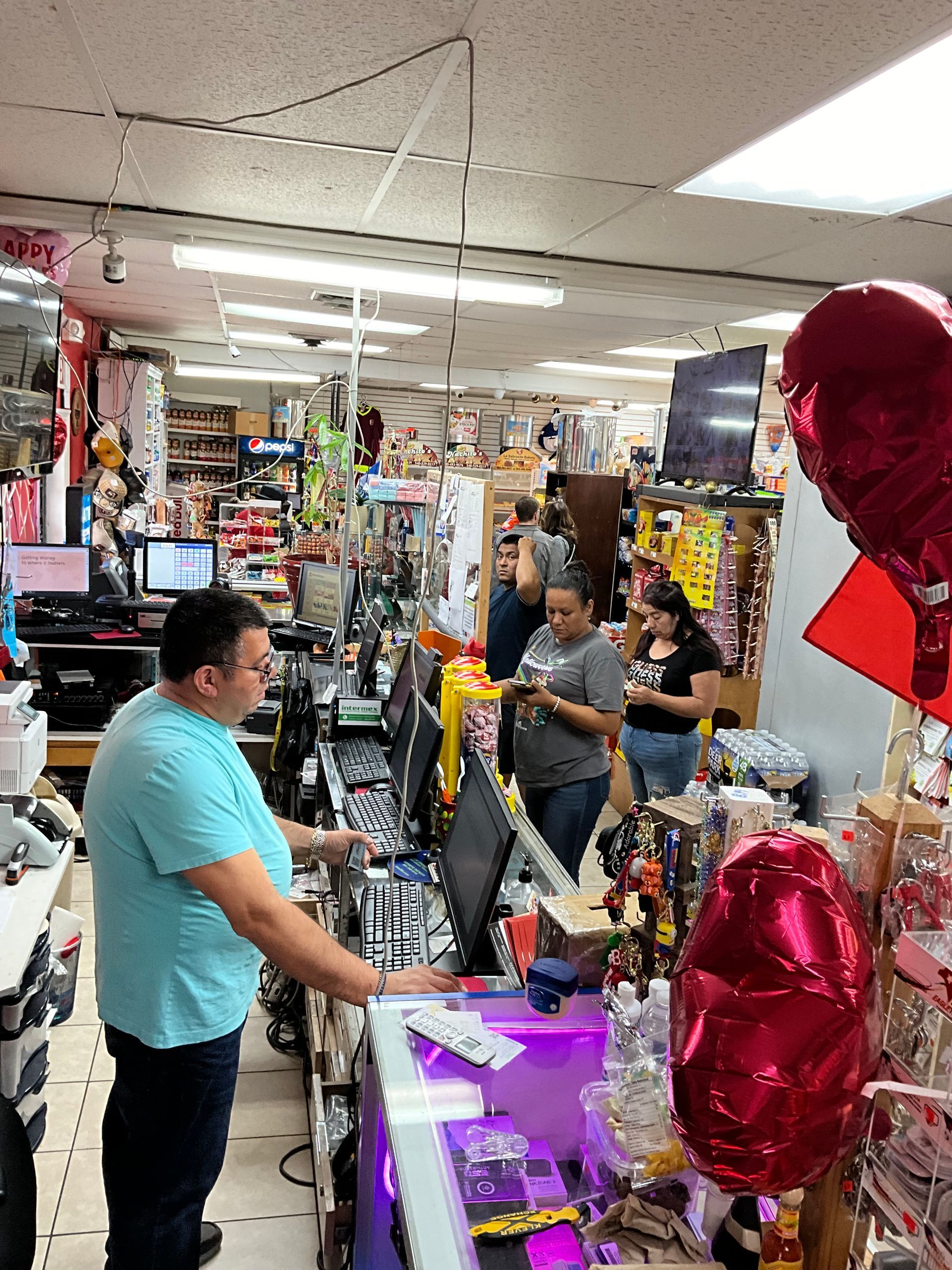 Man at a counter in a crowded store, customers browse merchandise, red heart balloons in the foreground.