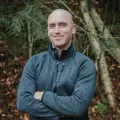 A man is standing in the woods with his arms crossed and smiling
