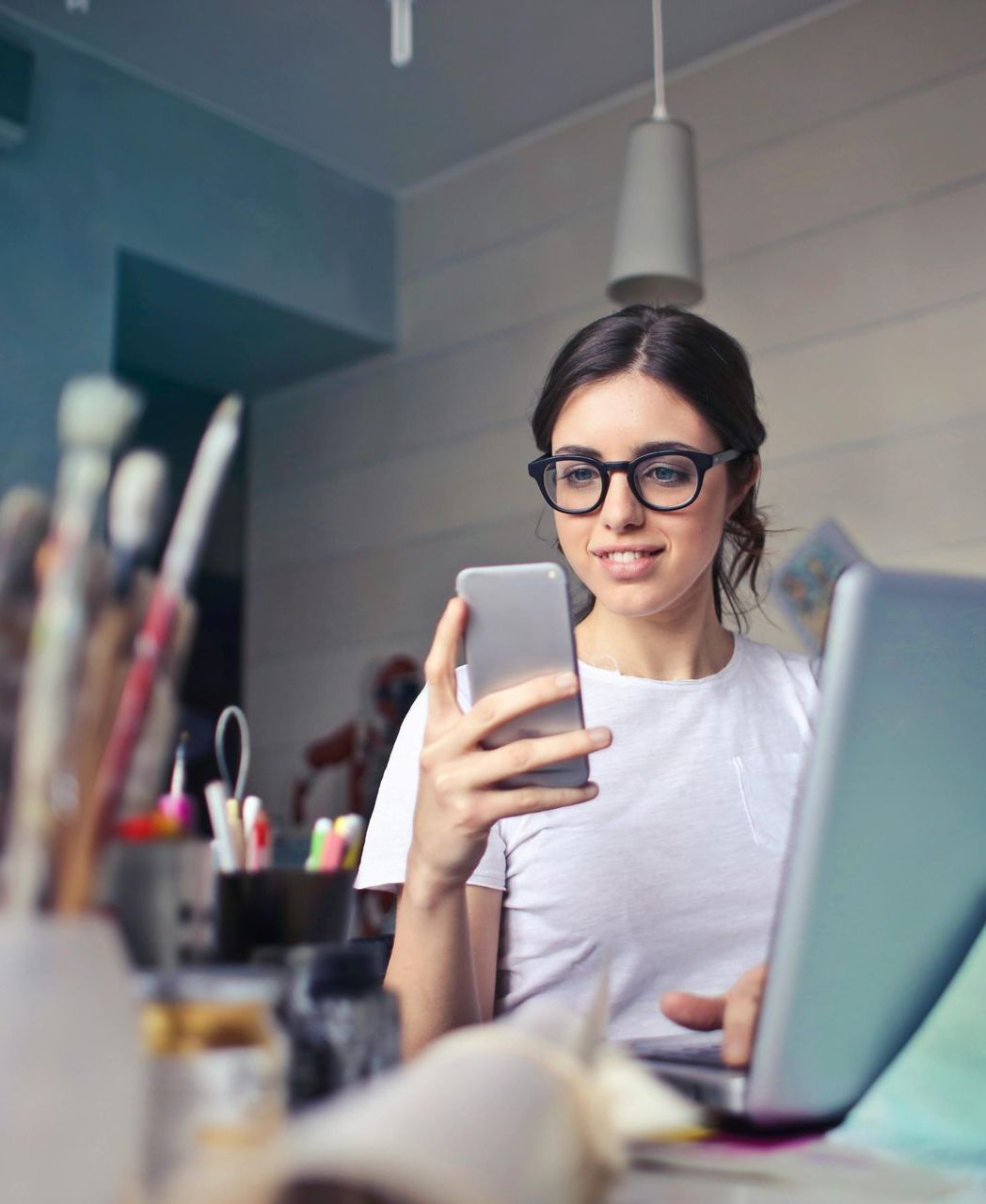 A woman is sitting in front of a laptop holding a cell phone.