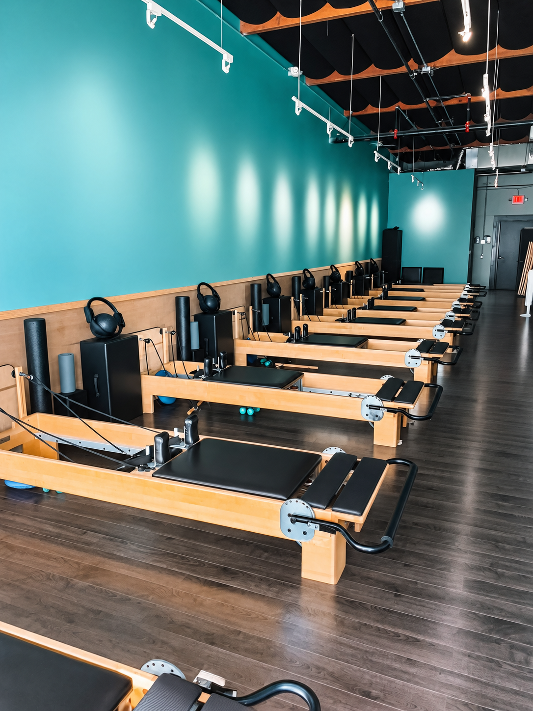 Pilates studio with reformer machines in a row, teal wall and dark wood floor.
