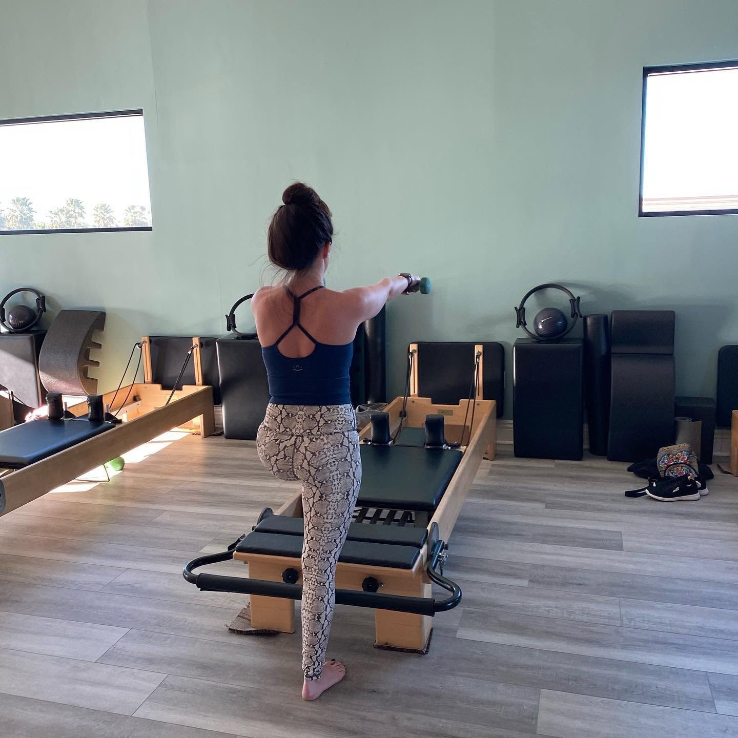 Person on a Pilates reformer in a studio, viewed from behind, with large windows and exercise equipment.