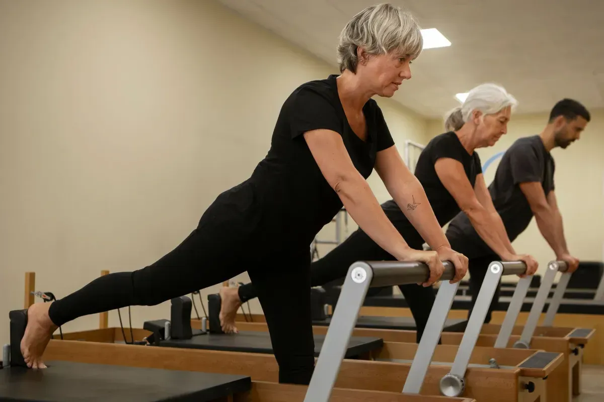 Person on a Pilates reformer in a studio, viewed from behind, with large windows and exercise equipment.