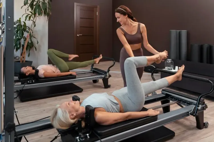 Pilates studio with two women exercising on reformers, one lying down while an instructor assists her leg stretch