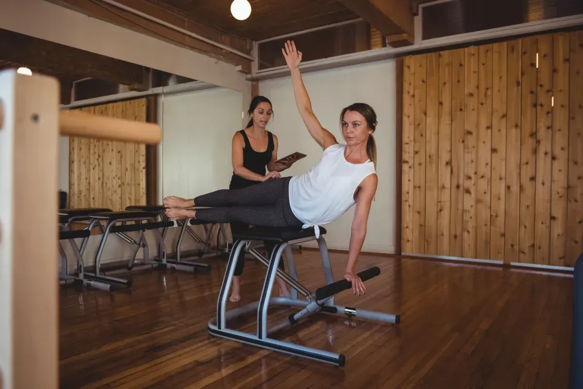 Woman balancing on one leg in a Pilates studio, holding a barre with outstretched arms.