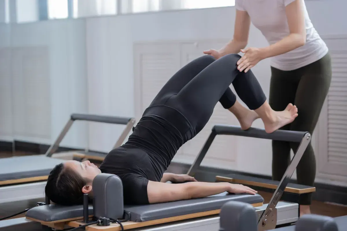 Woman balancing on one leg in a Pilates studio, holding a barre with outstretched arms.