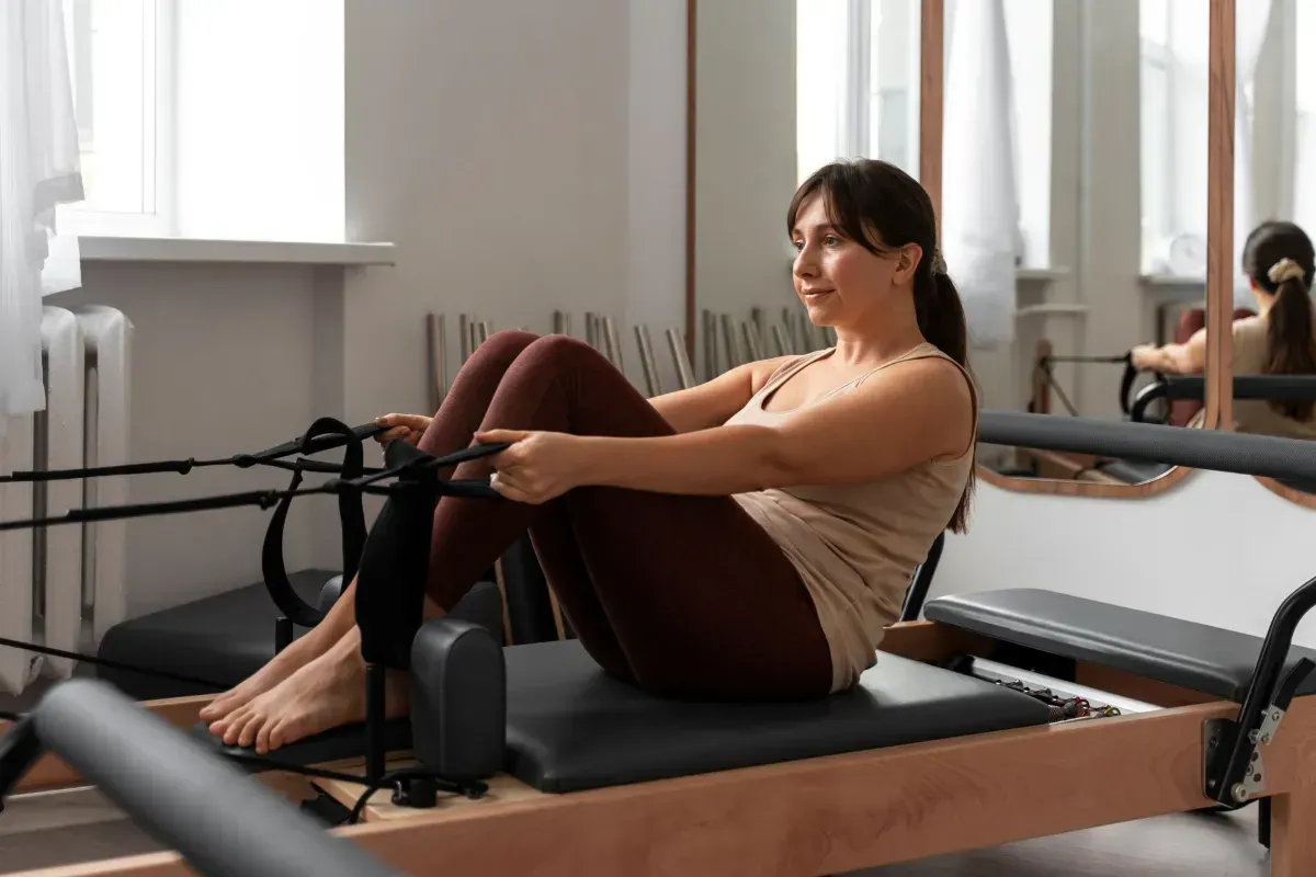 Person on a Pilates reformer in a studio, viewed from behind, with large windows and exercise equipment.