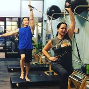Two people balancing on one leg in a gym, arms raised during a workout.