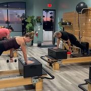People exercising on reformer Pilates machines in a bright studio with mirrors and wood flooring