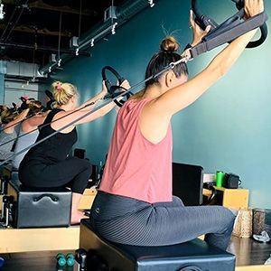 People doing Pilates on reformer machines in a bright studio, stretching arms overhead.