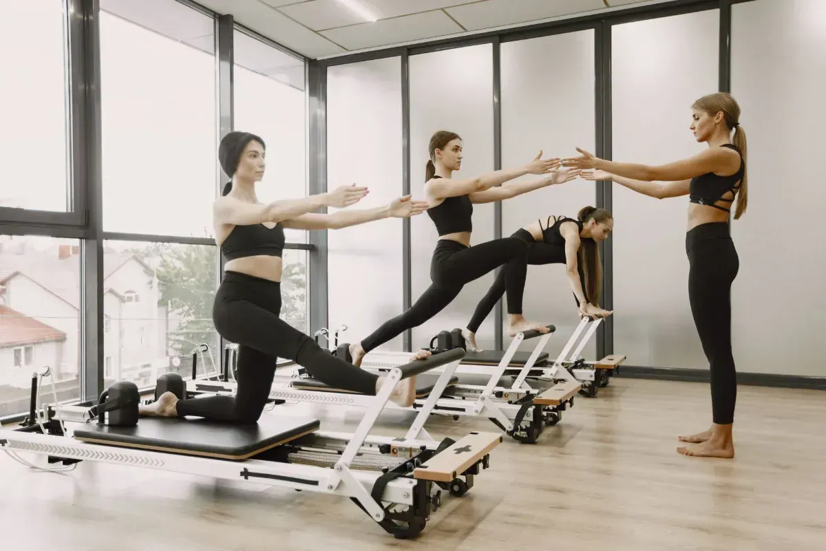 Three people exercising on reformer Pilates machines in a bright studio, stretching toward an instructor.