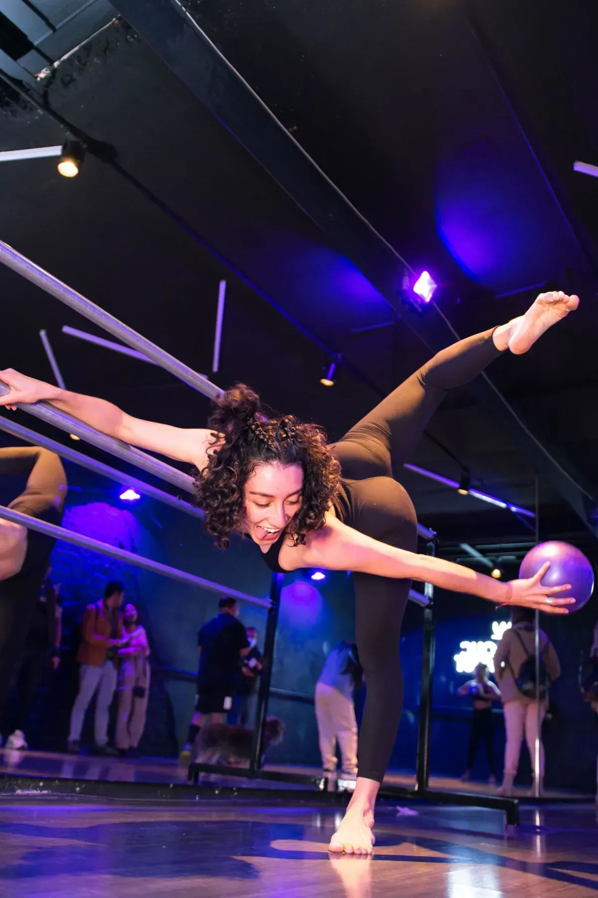 Dancer in a black costume balances in an extreme split pose under purple stage lights.