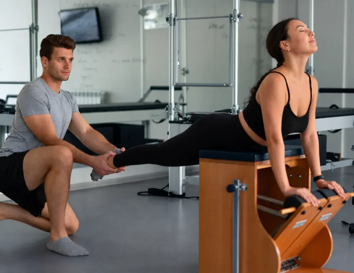Person on Pilates chair while instructor supports leg stretch in a studio