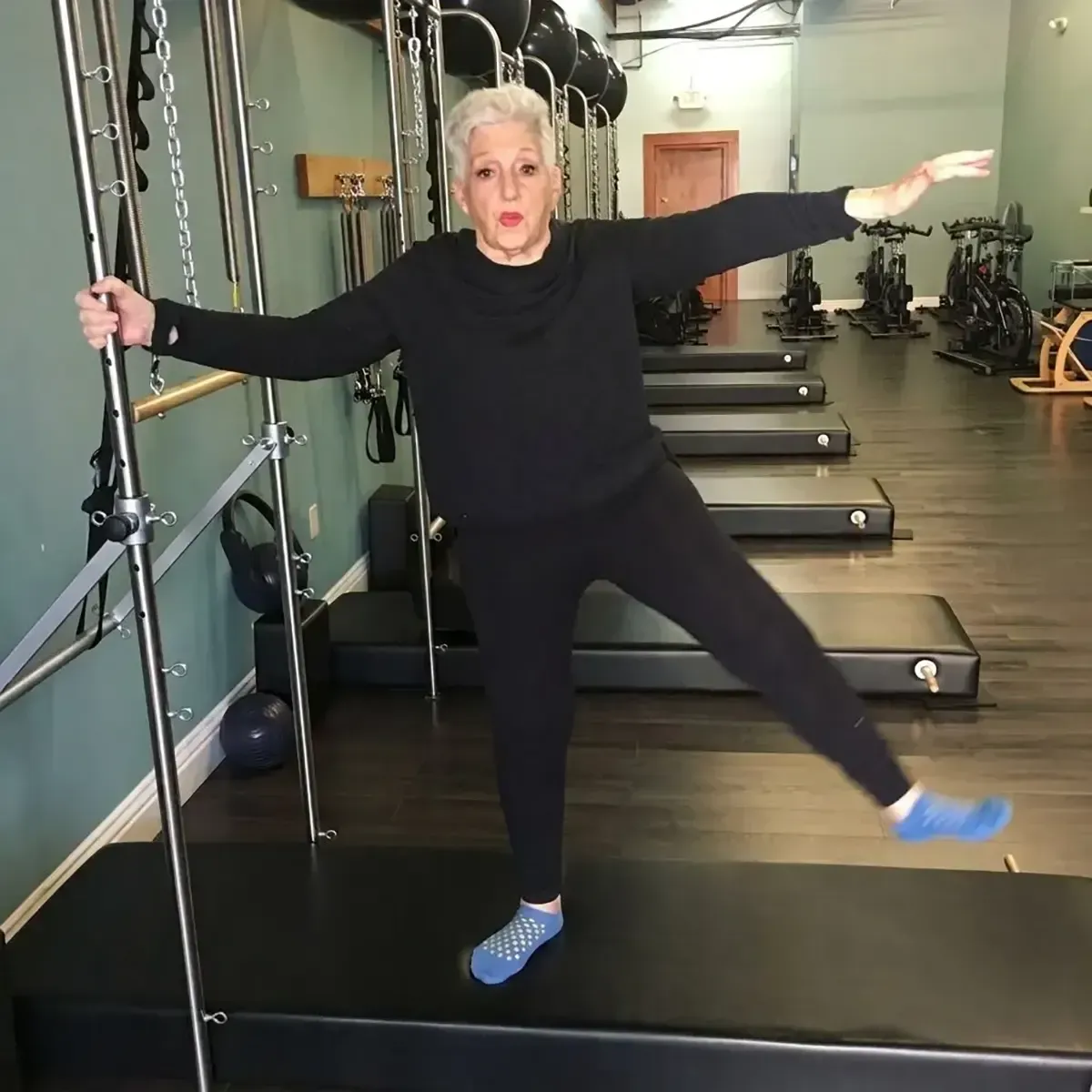 Woman balancing on one leg in a Pilates studio, holding a barre with outstretched arms.